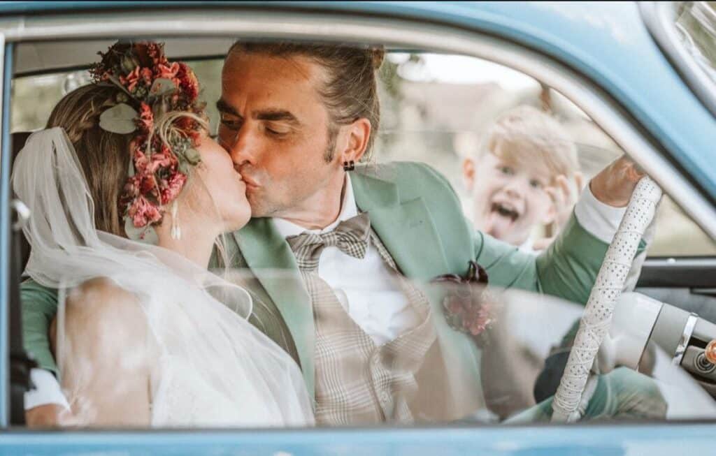 Mona und Daniel küssen sich in einem Auto, während ein Kind im Hintergrund lacht